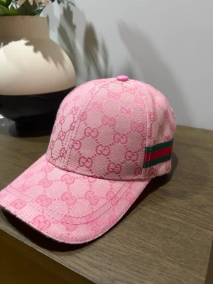 Rosa keps från Gucci med GG-mönster - Säljer en rosa keps från Gucci med det klassiska GG-monogrammet över hela kepsen. På sidan finns en grön och röd rand som ger extra Gucci-vibe. Kepsen har böjd skärm och är tillverkad i ett slitstarkt tyg, perfekt för dig som vill sticka ut.