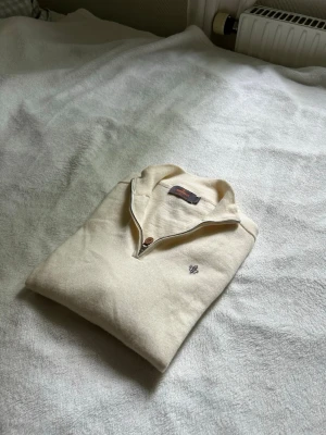 Beige Merino halvzip från Morris - Snygg beige stickad tröja från Morris med halv dragkedja och liten broderad logga på bröstet. Tröjan har en klassisk krage och är perfekt för lager-på-lager-stil. Materialet känns mjukt och bekvämt.