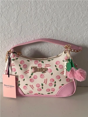 Juicy Couture rosa körsbärs-handväska - Söt handväska från Juicy Couture i vitt och ljusrosa med körsbärsmönster och guldfärgade detaljer. Väskan har en axelrem i rosa och kedja, samt en dekorativ pompom och löv. Perfekt för dig som gillar färgglada accessoarer och trendig design.