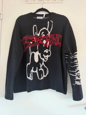 Svart stickad tröja med coolt motiv - Cool svart stickad tröja från Weekday med stort vitt motiv framtill och texten 'BE KIND' i rött över bröstet. Ena ärmen har ett vitt grafiskt mönster. Tröjan har rund hals och avslappnad passform, perfekt för en trendig streetwear-look. Aldrig använd, köpt på vinted
