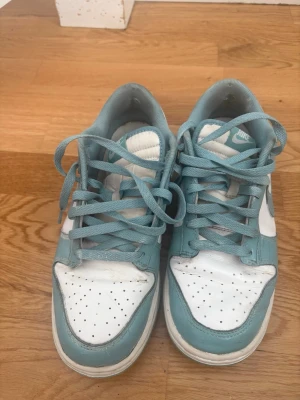 Nike Dunk Low blå och vita sneakers - Nike Dunk Low sneakers i ljusblått och vitt läder med klassisk swoosh på sidan. Skorna har perforerad tåbox, matchande blå skosnören och en låg siluett som ger en clean och sportig vibe. Perfekta för dig som gillar streetwear och vill sticka ut.