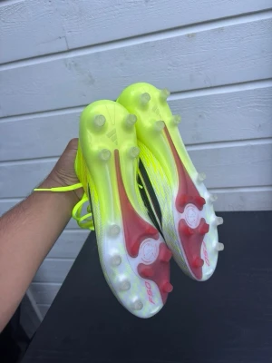 Neongula Adidas F50 fotbollsskor elite - Säljer ett par Adidas F50 fotbollsskor i neongult och vitt med svarta detaljer. Skorna har snörning, låg profil och mönstrad ovandel. Sulan har fasta dobbar i plast och inslag av rött. Perfekta för snabba spelare som vill sticka ut på planen.