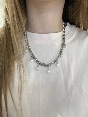 Silverfärgat halsband med pärlor - Säljer ett chunky silverfärgat halsband med hängande vita pärlor. Kedjan är grov och pärlorna är droppformade och sitter jämnt fördelade längs halsbandet. Perfekt för dig som vill ha en trendig accessoar med lite extra edge. Handgjort och rostfritt stål