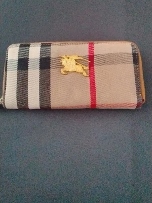 Rutig plånbok med gulddetalj - Snygg plånbok med klassiskt rutigt mönster i beige, svart, vitt och rött. Plånboken har en guldfärgad detalj med en riddare på häst framtill. Dragkedja runt om och stilren design, perfekt för att hålla koll på kort och cash.
