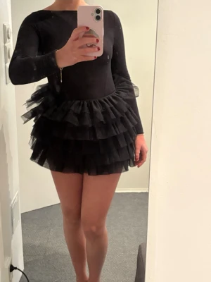 Svart volangklänning med lång ärm - Supersnygg svart kortklänning med långa ärmar och flera lager volanger i kjolen. Klänningen har en tight passform upptill och en fluffig kjol i tyll som ger en cool och trendig look. Perfekt för dig som vill sticka ut och älskar volym!