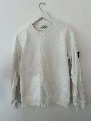Vit sweatshirt från Stone Island - Snygg vit sweatshirt från Stone Island med klassisk rund hals och ribbade muddar. Tröjan har en diskret patch på ärmen och är tillverkad i mjuk bomull för maximal komfort. Perfekt för en clean och stilren look.