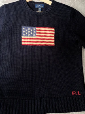 Mörkblå stickad tröja Polo Ralph Lauren - Mörkblå stickad tröja från Polo Ralph Lauren med amerikansk flagga broderad på bröstet och RL-broderi nertill. Tröjan har rund halsringning, långa ärmar och ribbstickade muddar. Perfekt för dig som gillar klassisk preppy stil.
