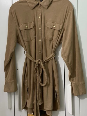 Beige skjortjumpsuit med knytskärp - Stilren beige skjortjumpsuit med långa ärmar, knappar framtill och två bröstfickor med lock. Tillverkad i mjukt tyg och har ett matchande knytskärp i midjan för en snygg siluett. Perfekt för dig som gillar en avslappnad men trendig look.