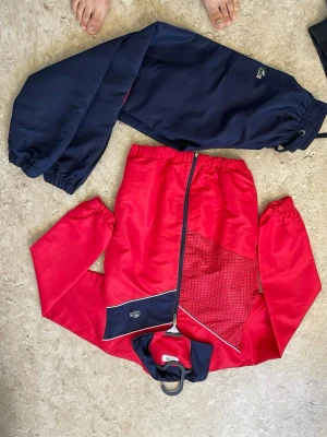 Röd och marinblå träningsdress - Snygg träningsdress från Lacoste med röd jacka och marinblå detaljer samt matchande marinblå byxor. Jackan har hel dragkedja, rutmönster på ena sidan och Lacoste-logga på bröstet. Byxorna har resår i midja och benslut. Perfekt för sport eller chill.