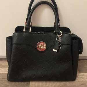 Svart handväska från Guess - Svart handväska från Guess med präglat mönster och guldfärgade detaljer. Väskan har dubbla handtag, dragkedja upptill, ytterfack med dragkedja och flera innerfack. Axelband medföljer till väskan. Se sista bilden för ytterst liten defekt som inte påverkar dess funktion. Väskan är i sin helhet i fint skick. 