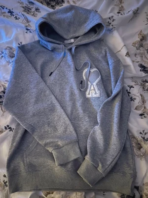 Ami Paris Hoodie - Säljer en grå hoodie från AMI Paris med stor broderad A-logga på bröstet. Hoodien har huva med dragsko, magficka och ribbade muddar vid ärmar och nederkant. Perfekt för en chill och stilren look. Tröjan är i storleken M men passar också bra S.