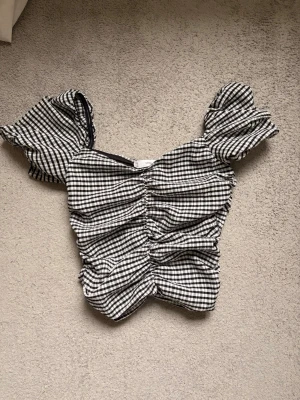 Rutig puffärmad topp i svartvitt - Supersöt topp med svartvitt rutigt mönster och puffiga korta ärmar. Toppen har rynkade detaljer framtill som ger en snygg formad look. Toppen är ifrån Mango och är i storlek EUR M med en dragkedja på sidan. Jag säljer den då den är för liten för mig :)