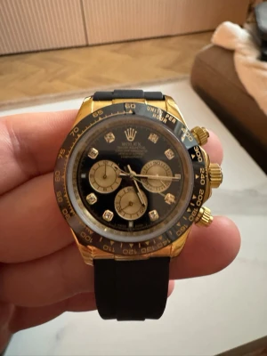 Role Daytona guld/svart kronograf - Snygg Role Daytona kronograf med svart urtavla, guldfärgade detaljer och svarta subdials. Boetten är i guld och armbandet är svart, troligtvis i syntetmaterial. Urtavlan har diamantindex och tre mindre ur för tidtagning. Automatisk är den. Justerbar. 