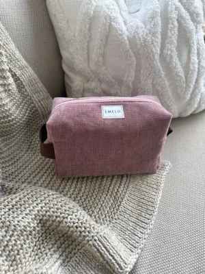 Rosa necessär från Emelo - Söt och stilren necessär i dammigt rosa tyg från Emelo. Väskan har en rektangulär form, dragkedja upptill och ett brunt handtag på sidan. Perfekt för smink eller småprylar. Materialet är tyg med en mjuk känsla och enkel design. Storlek medium. Mått ca 20x12x8 cm
