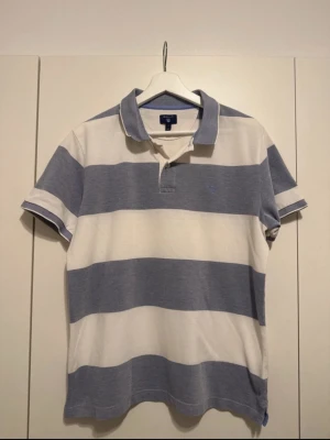 Randig blåvit t-shirt från Gant - Snygg t-shirt från Gant med breda horisontella ränder i ljusblått och vitt. Klassisk pikékrage, korta ärmar och knappslå framtill. Tillverkad i mjuk bomull som känns skön mot huden. Perfekt för en clean och avslappnad stil.