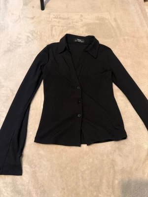 Svart ribbad skjorta med knappar - Stilren svart skjorta med ribbad struktur och långa ärmar. Skjortan har klassisk krage, knäppning framtill och en liten slits vid ärmslutet för extra detalj. Perfekt för en clean och modern look.