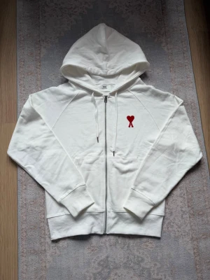 Vit zip hoodie från Ami Paris - Säljer en clean vit hoodie från Ami Paris med dragkedja och huva. På bröstet finns Ami:s ikoniska röda hjärta med bokstaven A. Hoodien har dragsko i huvan och känguruficka framtill. Tillverkad i mjuk bomull för skön känsla.