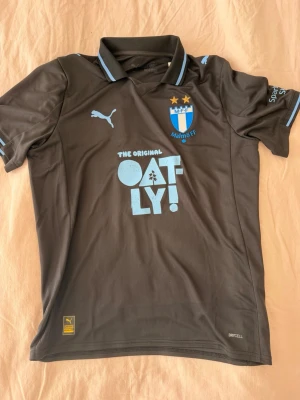 Malmö FF bortatröja Puma grå - Säljer en grå Malmö FF bortatröja från Puma med ljusblå detaljer och klubbmärke på bröstet. Tröjan har sponsorloggor från Oatly och Akea, korta ärmar och klassisk krage. Tillverkad i funktionsmaterial för träning och match.