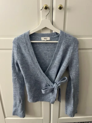 Ljusblå omlottkofta  - Söt ljusblå stickad omlottkofta med knytband i sidan och djup v-ringning. Koftan har långa ärmar och ribbade muddar vid ärmslut och nederkant. Perfekt för lager på lager och en chill look.