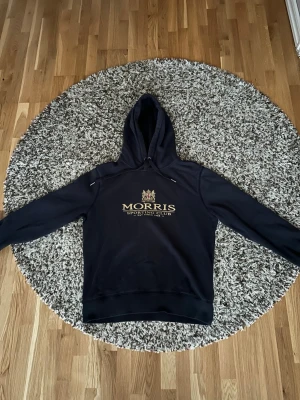 Svart hoodie från Morris - Snygg svart hoodie från Morris med guldfärgat tryck och texten 'Morris Sporting Club' på bröstet. Klassisk huva med dragsko och ribbade muddar vid ärmar och nederkant. Perfekt för en avslappnad och stilren look.