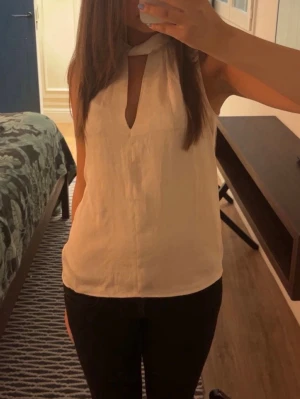 Vit ärmlös blus med knyt i halsen - Stilren vit ärmlös blus med snygg knytning i halsen och en djup V-ringning framtill. Blusen har en lätt och luftig känsla, perfekt till sommaren. Materialet ser ut att vara ett tunt, mjukt tyg som faller fint.