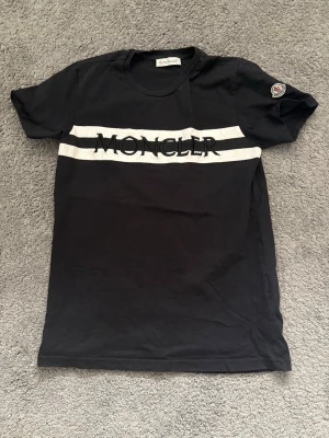 Svart Moncler t-shirt med logga - Snygg svart t-shirt från Moncler med vit rand och stor Moncler-logga över bröstet. Klassisk rund halsringning och korta ärmar. Tillverkad i mjuk bomull för skön känsla. Perfekt för dig som gillar stilrena och exklusiva plagg.