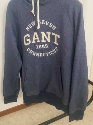 Blå hoodie från GANT med tryck - Mörkblå hoodie från GANT med vit text 'NEW HAVEN GANT 1949 CONNECTICUT' på bröstet. Klassisk känguruficka framtill, ribbade muddar och justerbar huva med vita snören. Mjuk bomullsblandning som känns skön mot huden.