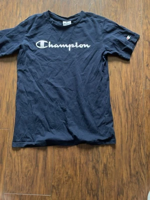 Svart Champion t-shirt med logga - Snygg svart t-shirt från Champion med stor vit logga över bröstet och det klassiska Champion-märket på ärmen. T-shirten har rund hals och korta ärmar, perfekt för en avslappnad och sportig stil.