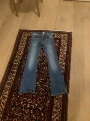 Blå bootcut jeans från Gina Tricot - Säljer ett par blå bootcut jeans från Gina Tricot med snygg slitning på låren och dubbla knappar i midjan. Klassisk femficksmodell i stretchigt denimtyg som sitter skönt och ger en avslappnad look.
