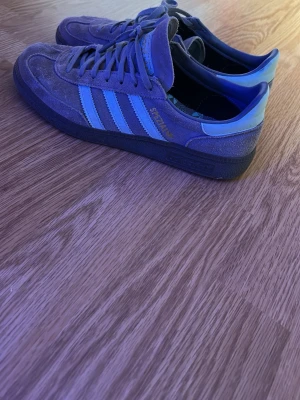 Adidas Samba sneakers blå/grå mocka - Snygga Adidas Samba sneakers i blått och grått mockamaterial med klassiska tre ränder i ljusblått på sidan. Låg modell med svart sula och guldfärgad Samba-text. Perfekta för dig som gillar retrostil och streetwear.