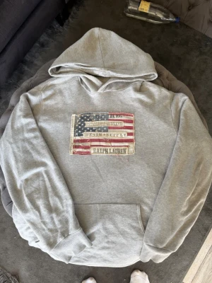 Grå hoodie med USA-flagga Ralph Lauren - Säljer en grå hoodie från Ralph Lauren med ett stort USA-flagga patch på bröstet och texten 'DENIM & SUPPLY'. Hoodien har huva och en stor magficka. Perfekt för dig som gillar klassisk streetstyle och vill ha något bekvämt och snyggt.