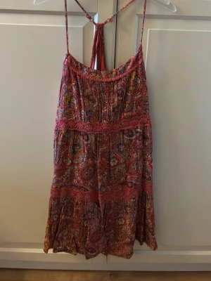 Mönstrad sommarklänning med spetsdetaljer - Säljer en bohemisk kortklänning med tunna axelband och färgglatt paisleymönster i rosa, rött och inslag av blått och gult. Klänningen har fina spetsdetaljer och volangkant upptill. Storlek M. I fint skick. Från Hollister. 