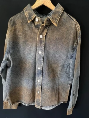 Tvättad grå jeansjacka unisex - Snygg jeansjacka i tvättad grå färg med klassisk krage och vita knappar framtill. Jackan har avslappnad passform, långa ärmar med knapp vid ärmslut och en rak nederkant. Perfekt för dig som gillar en vintage och avslappnad stil.