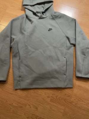 Grå Nike tech hoodie - Snygg grå hoodie från Nike med minimalistisk design och svart Nike-logga på bröstet. Hoodien har en justerbar huva med dragsko och långa ärmar. Tillverkad i mjukt material som känns skönt mot huden. Perfekt för en avslappnad streetwear-look. Det ör storlek XL men passar mycket bättre till dig som har strl L😊