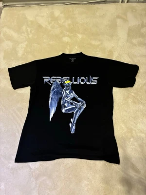 Svart Rebellious t-shirt med tryck - Cool svart t-shirt från Rebellious med ett futuristiskt tryck av en ängel i silver och blå toner på framsidan. T-shirten har klassisk passform och korta ärmar. Perfekt för dig som gillar statement-plagg och grafiska motiv.