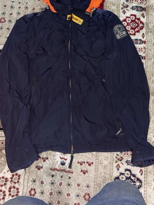 Mörkblå vindjacka från Parajumpers - Snygg mörkblå vindjacka från Parajumpers med orange detaljer och huva. Jackan har dragkedja framtill, två sidofickor med dragkedja och meshfoder på insidan. Märkespatch på ärmen och justerbar nederkant. Perfekt för blåsiga dagar.
