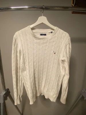 Vit kabelstickad tröja från GANT - Kabelstickad vit tröja från GANT i premium bomull. Klassisk rund halsringning, ribbade muddar och liten GANT-logga broderad på bröstet. Perfekt för dig som gillar stilrena och tidlösa plagg.