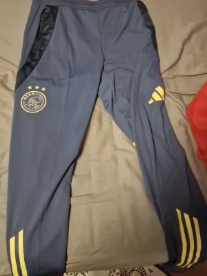 Blå Ajax träningsbyxor Adidas - Mörkblå träningsbyxor från Adidas med Ajax-logga och gula detaljer. Byxorna har resår i midjan, dragkedjefickor och gula ränder på benen. Tillverkade i ett lätt och stretchigt material som passar perfekt till träning eller sport.