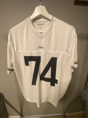 Vit Dallas sporttröja nr 74 - Vit sporttröja från New Garments med svart nummer 74 på framsidan och texten 'Dallas' ovanför. Tröjan har V-ringning, korta ärmar med svarta och bruna ränder samt ett Dallas-märke på ena ärmen. Tillverkad i ett ventilerande, mesh-liknande material