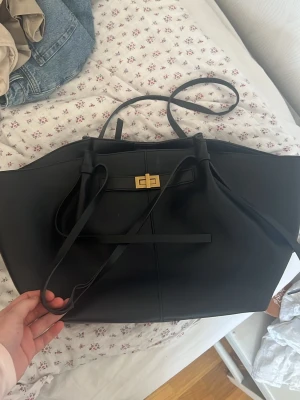 Svart shoppingväska med gulddetalj - Stilren svart shoppingväska i slätt konstläder med långa handtag och snygg guldfärgad vridlåsdetalj framtill. Väskan har en rymlig form och minimalistisk design, perfekt för att bära allt du behöver. Passar till många olika outfits och är enkel att matcha.