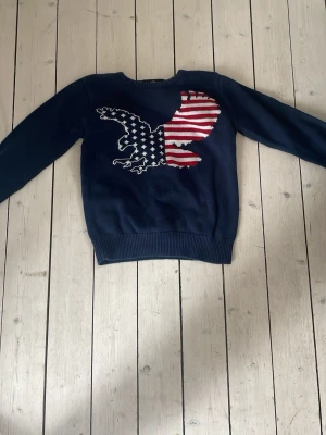 Grisch stickad tröja med amerikansk örn. - Mörkblå stickad tröja med en stor örn på bröstet, där örnen har ett amerikanskt flaggmönster i rött, vitt och blått. Tröjan har rund halsringning och ribbade muddar vid ärmslut och nederkant. Perfekt för dig som gillar coola statement-plagg.