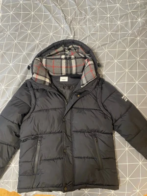 Svart pufferjacka från burberry - Svart pufferjacka från burberry med avtagbar huva och rutigt foder i grått, rött och vitt. Jackan har två dragkedjeförsedda fickor framtill och logotyp på ärmen. Perfekt för lite kyligare dagar och riktigt snygg att bära.