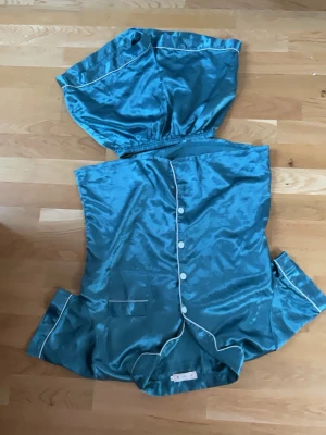 Turkos satinpyjamas med kort ärm - Snygg turkos pyjamas i glansigt satinmaterial med kortärmad skjorta och matchande shorts. Skjortan har vita knappar, kontrasterande vita kanter och en bröstficka. Perfekt för mysiga kvällar hemma.