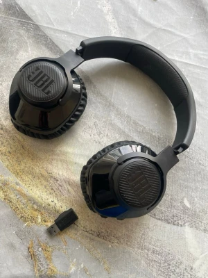 Svarta trådlösa hörlurar från JBL - Snygga svarta over-ear hörlurar från JBL med vadderad bygel och mjuka öronkuddar i konstläder. Hörlurarna är trådlösa och kommer med en USB-dongel för enkel anslutning till dator eller konsol. Perfekta för gaming, musik eller film.