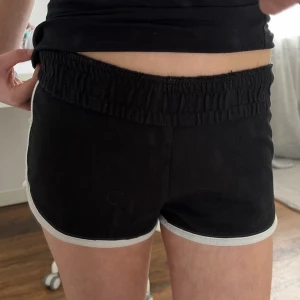 Svarta shorts med vit kant - Säljer ett par svarta shorts med vit kant och elastisk midja. Klassisk sportig look med korta ben och mjukt material, perfekta för sommarens alla aktiviteter.