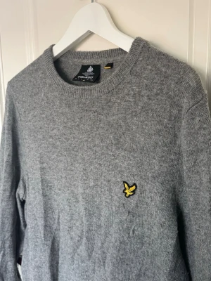 Grå stickad tröja från Lyle & Scott - Klassisk grå stickad tröja från Lyle & Scott med rund halsringning och den ikoniska gula örnloggan broderad på bröstet. Tröjan har ribbade muddar vid ärmslut och nederkant. Perfekt för lager på lager och enkel att matcha.