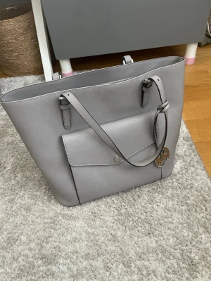 Grå toteväska från Michael Kors i äkta läder.(kom med prisförslag!!) - Stilren grå toteväska från Michael Kors i slätt läder. Väskan har långa axelband, en stor ytterficka med knappstängning och ett dekorativt MK-hänge i metall. Perfekt för dig som gillar enkel och klassisk design.