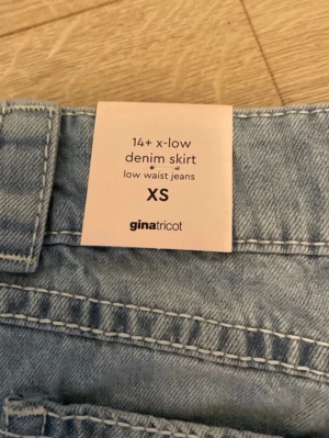 Ljusblå denimkjol från Gina Tricot - Ny med Prislapp. Säljer en ljusblå kort jeanskjol från Gina Tricot i storlek XS. Kjolen har låg midja, slitna detaljer och fransig nederkant. Klassisk femficksmodell med bälteshällor och knappstängning framtill. Perfekt för en trendig och avslappnad look.