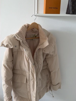 Beige pufferjacka med stor krage - Snygg beige pufferjacka från Gina Tricot med stor krage och dragkedja framtill. Jackan har praktiska fickor med lock och justerbar nederkant med dragsko. Perfekt för kalla vinterdagar och ger en trendig look.