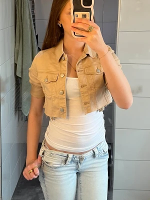 Beige croppad jeansjacka - Säljer en beige croppad jeansjacka med korta ärmar och klassisk krage. Jackan har två bröstfickor med lock och silverfärgade knappar framtill. Perfekt för dig som vill ha en trendig och unik twist på den klassiska jeansjackan.